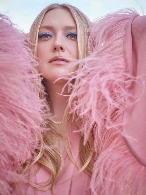 Close-up of blonde woman wearing a pink feathered jacket and blue eye makeup, looking directly at the camera with a neutral expression.