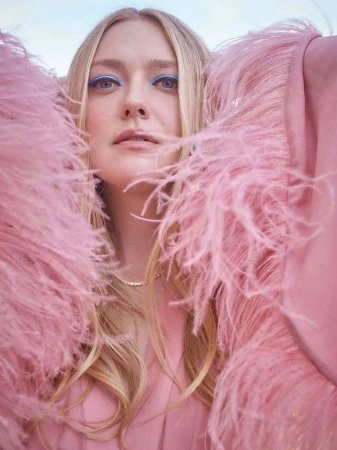 Close-up of blonde woman wearing a pink feathered jacket and blue eye makeup, looking directly at the camera with a neutral expression.
