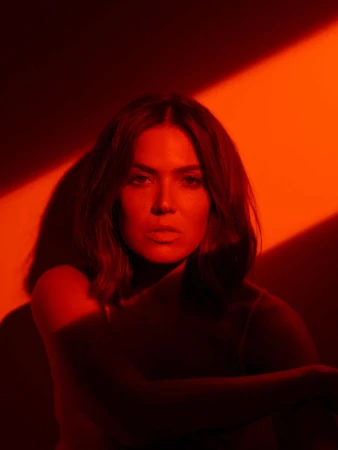 Close-up portrait of a woman bathed in dramatic red and orange light with sharp angular shadows on the wall behind her.