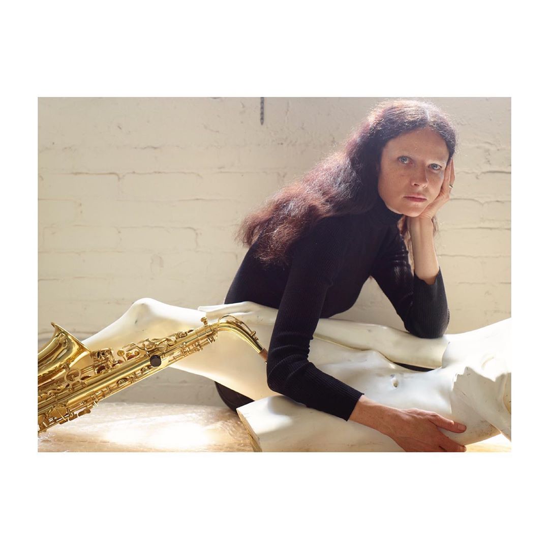 Woman with long brown hair in a black turtleneck posing with a saxophone draped over a white sculpture in a brightly lit room.