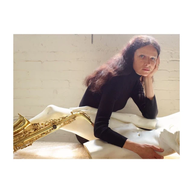 Woman with long brown hair in a black turtleneck posing with a saxophone draped over a white sculpture in a brightly lit room.