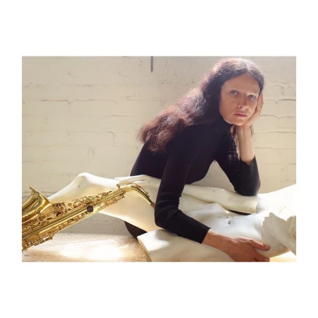 Woman with long brown hair in a black turtleneck posing with a saxophone draped over a white sculpture in a brightly lit room.