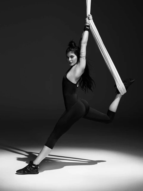 A woman in sportswear gracefully hangs on aerial silks in a black and white studio shot, lit from below, showcasing her strength and flexibility.