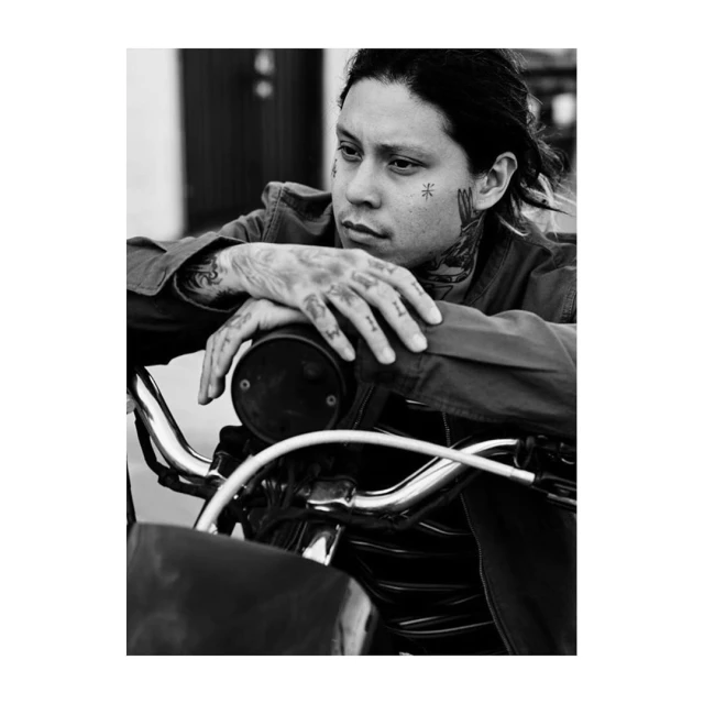 Monochrome portrait of tattooed man with serious expression leaning on motorcycle handlebars, gazing into the distance outdoors.