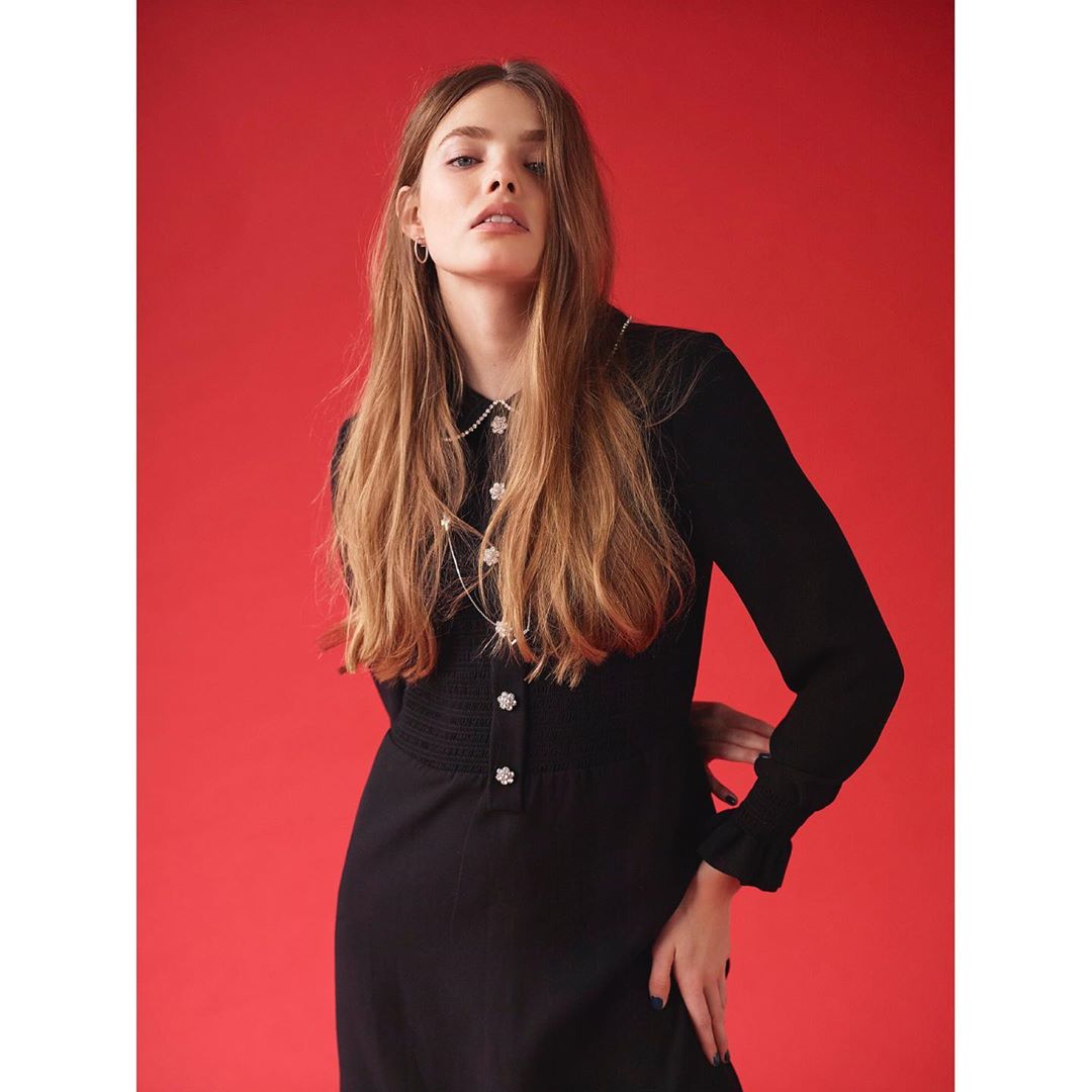 A stylish woman poses against a red background, wearing a long-sleeved black dress with pearl-like embellishments and jewelry, looking at the camera.