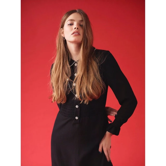 A stylish woman poses against a red background, wearing a long-sleeved black dress with pearl-like embellishments and jewelry, looking at the camera.