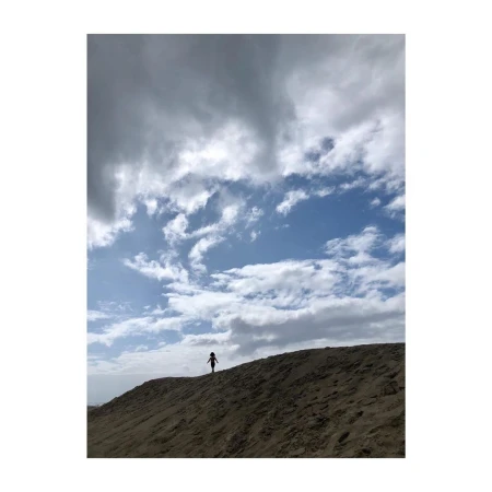 A small figure stands atop a sandy dune beneath a vast, partly cloudy sky captured in a vertical frame.