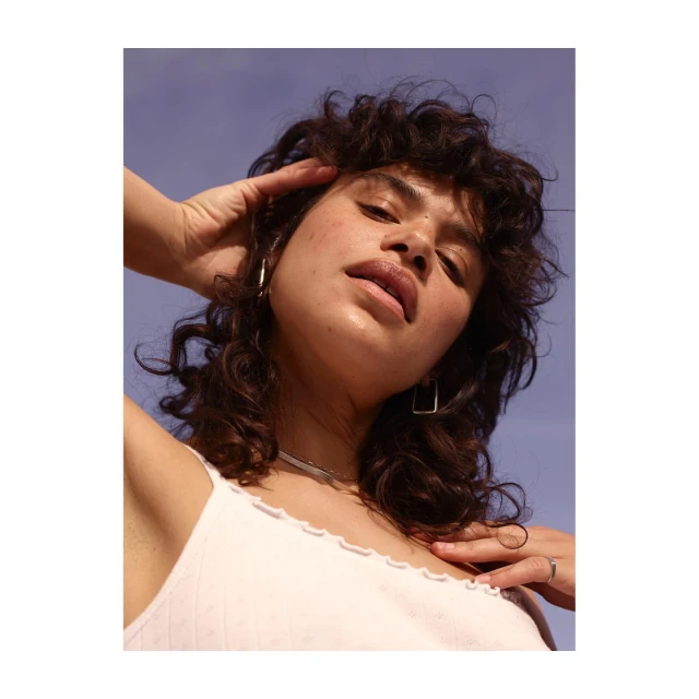 Portrait of a woman with curly brown hair, wearing a white top and silver jewelry, posing in front of a clear blue sky, shot from below.