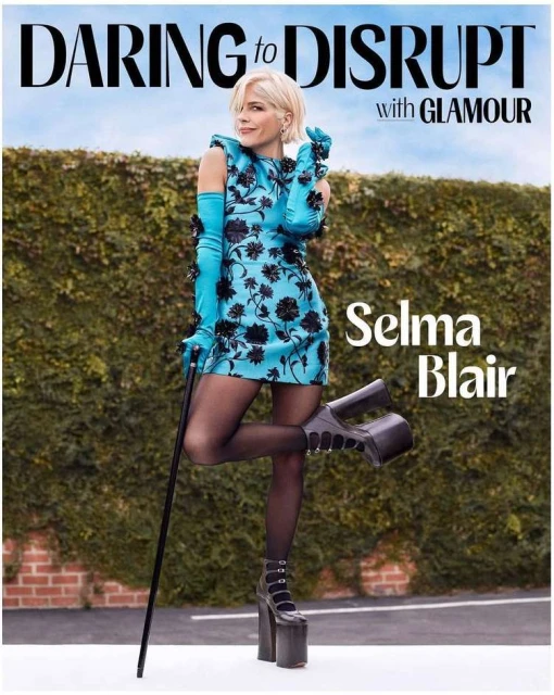Selma Blair poses in a blue floral dress, black stockings, and platform heels. She's holding a cane against a hedge and brick wall.
