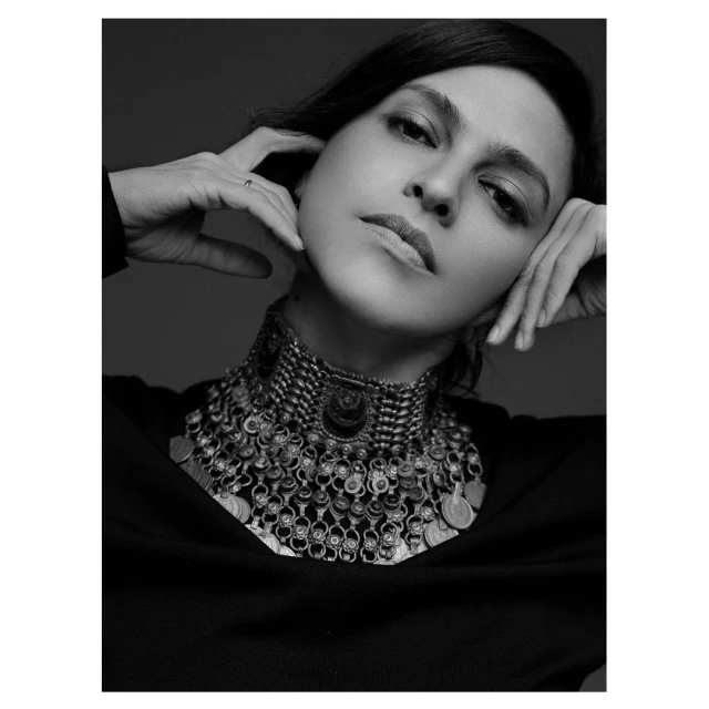 Monochrome portrait of a woman with dark hair, wearing a large, elaborate coin and metal necklace, hands near her face.