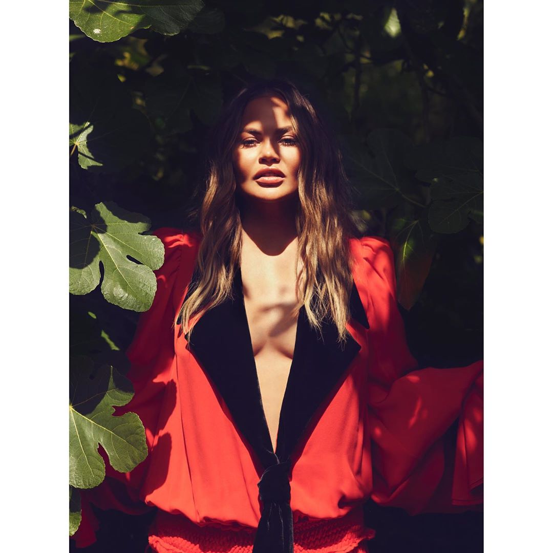 A woman with long wavy brown hair poses outdoors in a low-cut red top with black trim surrounded by green foliage in bright sunlight.
