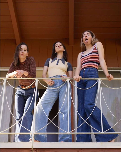Three young women in 1970s-style clothing pose on a balcony, wearing flared jeans, long-sleeved tops, and vintage accessories in outdoor light.