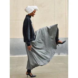 Woman wearing white turban, dark turtleneck sweater and long grey skirt kicking her leg up outdoors.