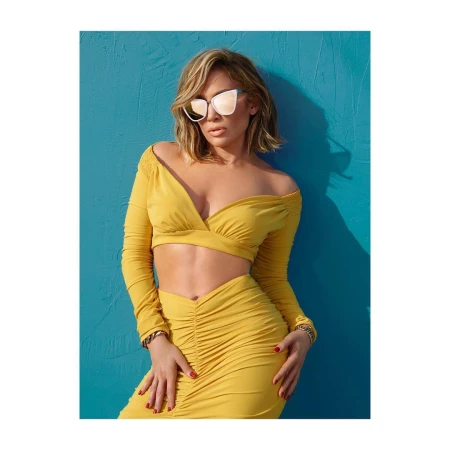Jennifer Lopez wearing a mustard yellow crop top and skirt with white sunglasses against a blue wall
