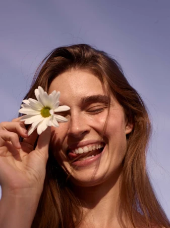 Close-up portrait of a smiling woman holding a white daisy flower in front of her eye against a clear sky background