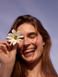 Close-up portrait of a smiling woman holding a white daisy flower in front of her eye against a clear sky background