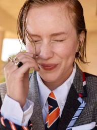 Close-up portrait of a woman with wet hair smiling with eyes closed, wearing a tweed jacket, white shirt, and striped tie.