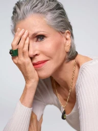 Close-up portrait of Jane Fonda with short grey hair, wearing a white ribbed top, a green ring, and a gold necklace.