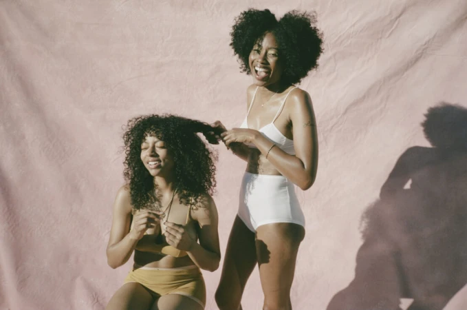 Two Black women with curly hair smiling against a pink backdrop, one standing in white underwear and the other seated wearing yellow underwear.