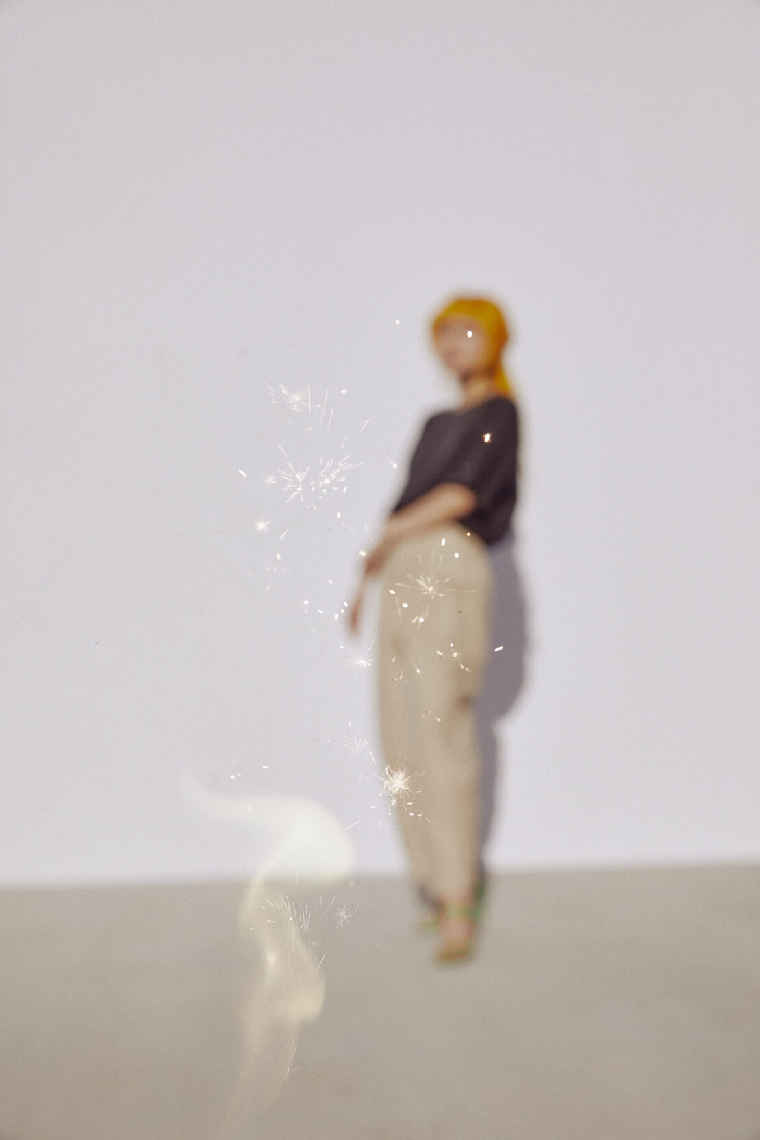 Woman with red hair and a dark top poses in front of a white wall with sparklers. Out of focus with shallow depth of field.