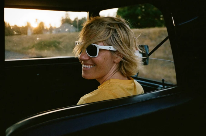 Blonde woman wearing white sunglasses and yellow t shirt smiling while looking back from inside a car