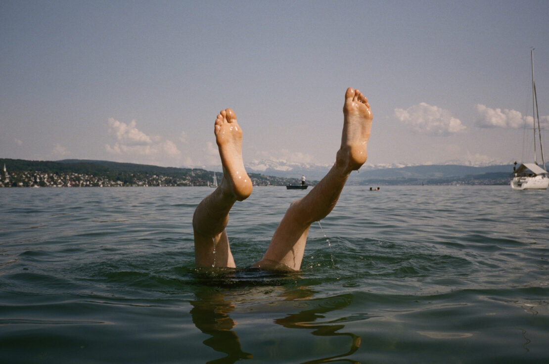 Person's legs and feet sticking out of rippled water on a sunny day, with a distant shoreline, buildings, and boat in the background.
