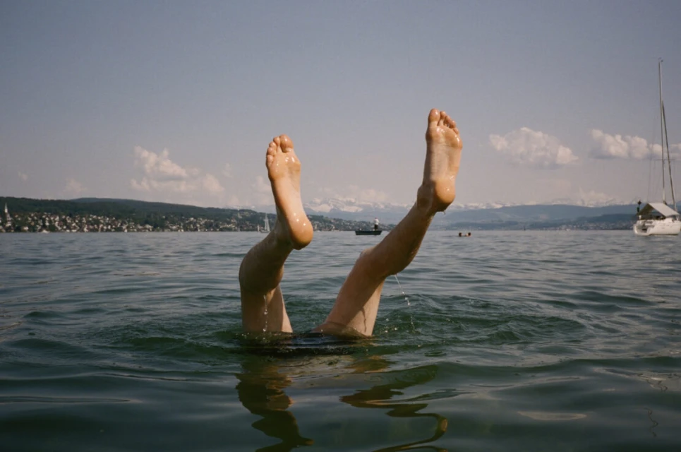 Person's legs and feet sticking out of rippled water on a sunny day, with a distant shoreline, buildings, and boat in the background.