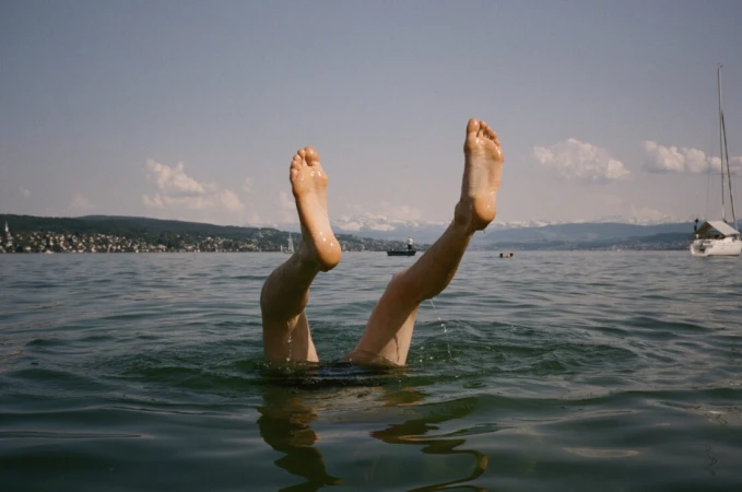 Person's legs and feet sticking out of rippled water on a sunny day, with a distant shoreline, buildings, and boat in the background.