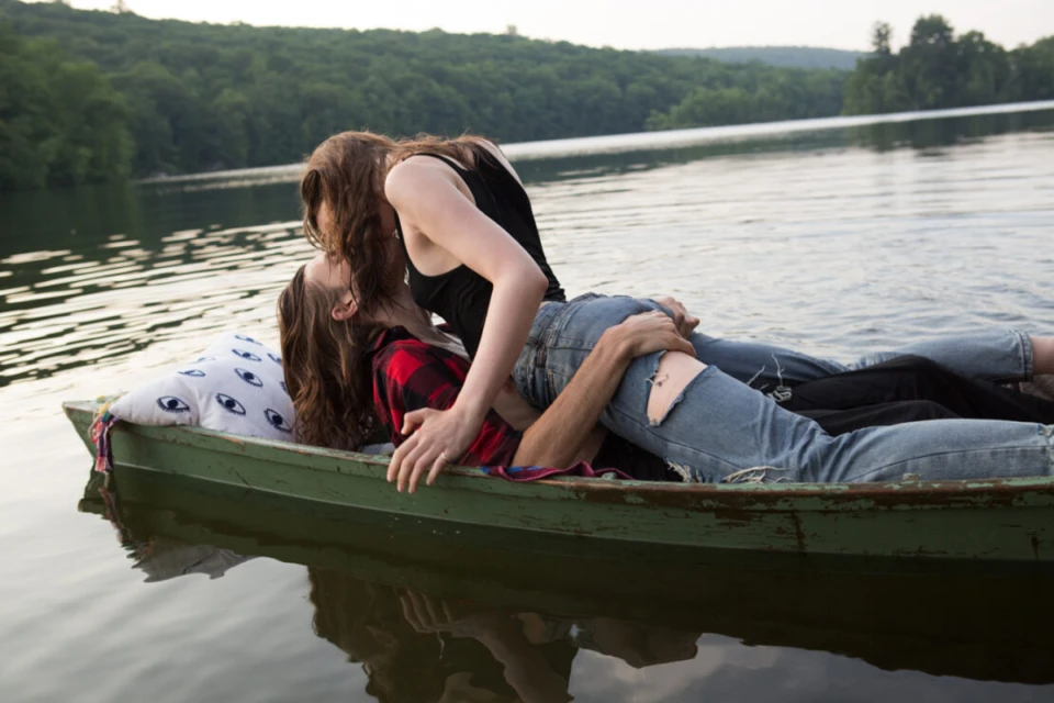 Two young adults embrace intimately in a green rowboat on a serene lake surrounded by lush green trees during daylight.