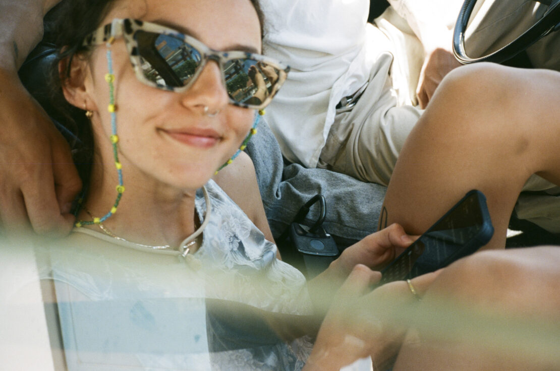 Smiling woman with patterned sunglasses and beaded chain poses inside a vehicle, holding a cell phone with a hand resting on her shoulder.