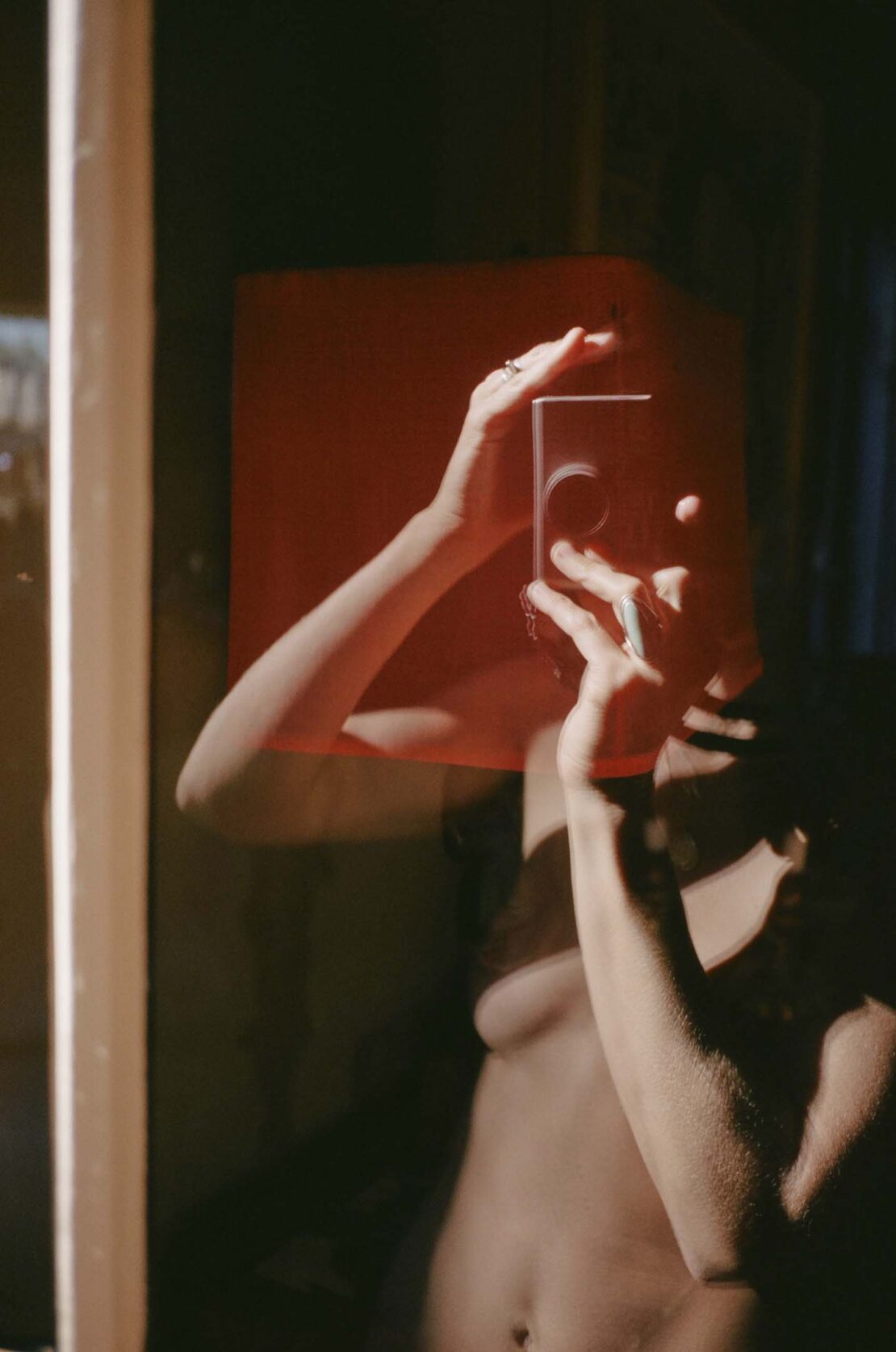 A partially nude woman takes a mirror selfie using a modern phone with a red block in the foreground against a shadowed interior.