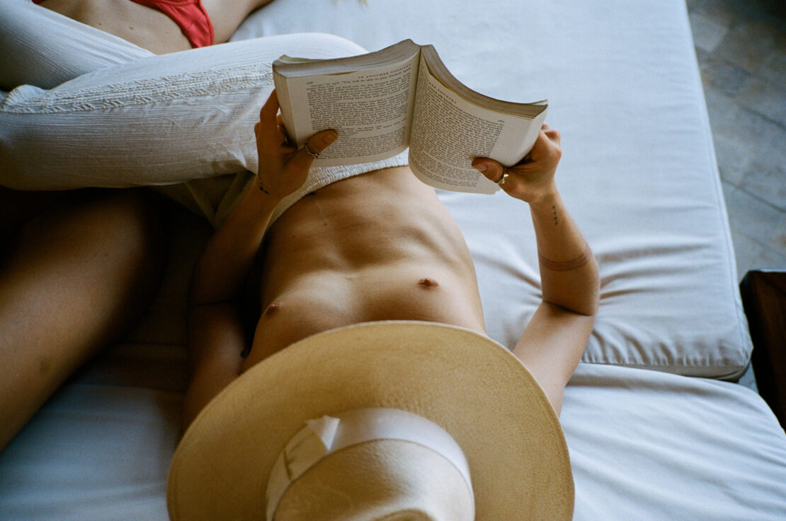 Person lying down on a white bed reading an open book while wearing a straw hat