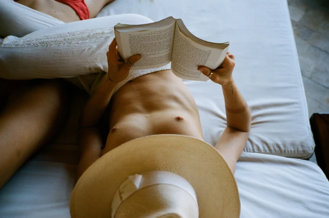 Person lying down on a white bed reading an open book while wearing a straw hat