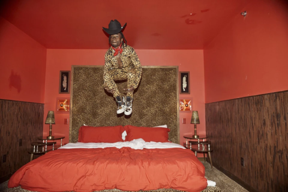 Person in a leopard-print suit and cowboy hat jumps over a bed in a bedroom with red walls, a leopard-print headboard, and patterned lamps.