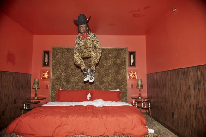 Person in a leopard-print suit and cowboy hat jumps over a bed in a bedroom with red walls, a leopard-print headboard, and patterned lamps.