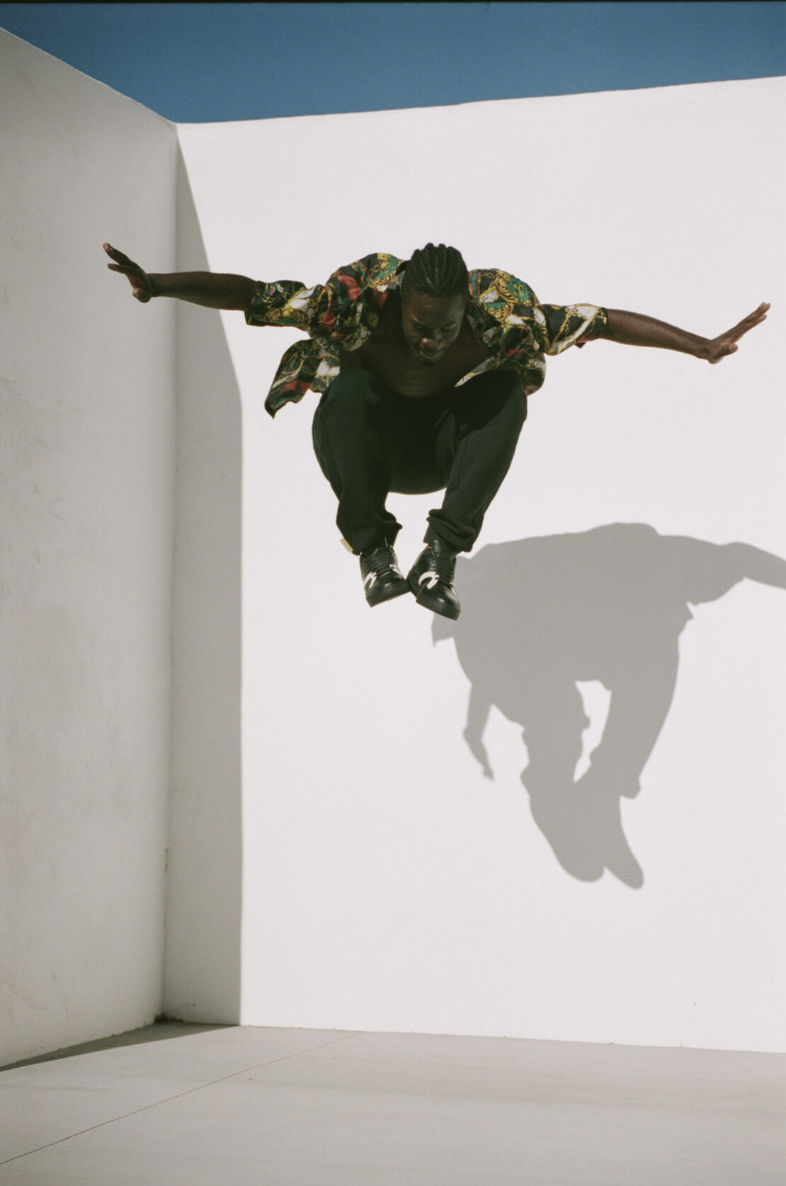 Athletic Black man in mid-air jump with arms outstretched, wearing floral shirt and black pants against white background with shadow.