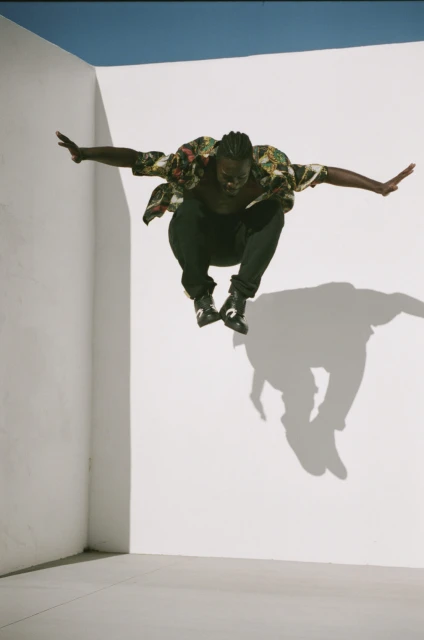 Athletic Black man in mid-air jump with arms outstretched, wearing floral shirt and black pants against white background with shadow.