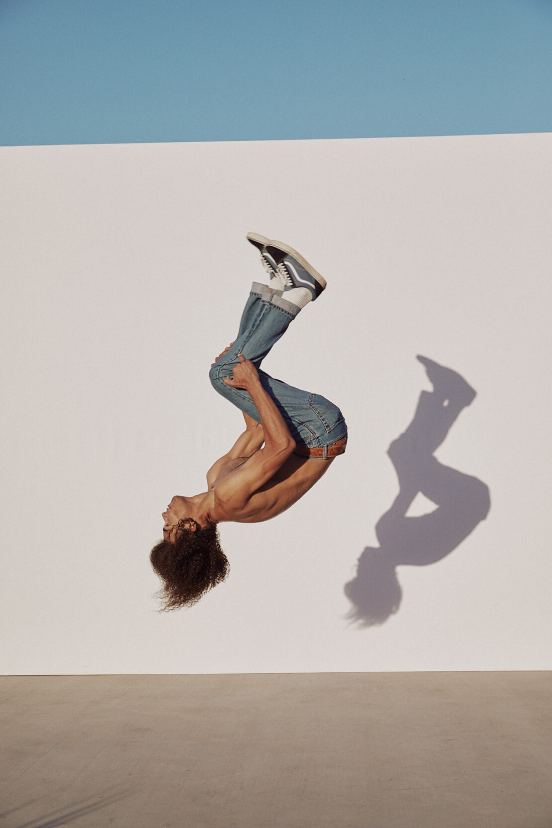 A male gymnast does a backflip against a white wall under a clear blue sky, showcasing strength and agility in a dynamic pose. 