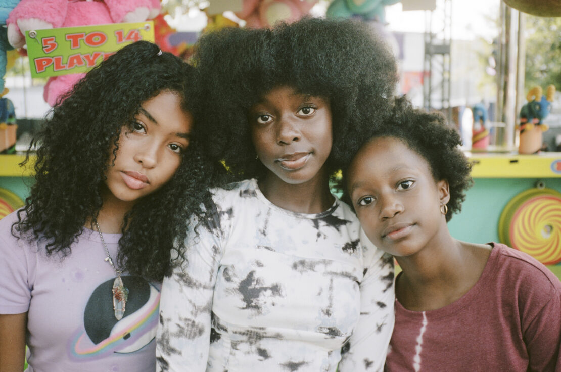 Three young women with natural hair pose outdoors. Each has a different hairstyle: curly, straight, and afro. All wear casual shirts.
