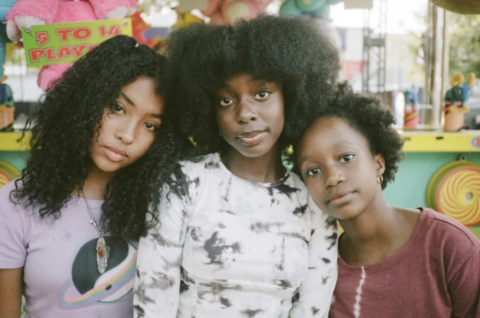 Three young women with natural hair pose outdoors. Each has a different hairstyle: curly, straight, and afro. All wear casual shirts.