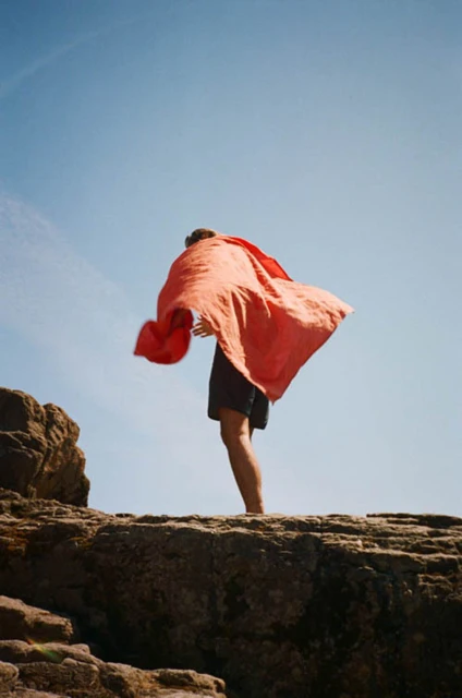 Man stands on rocky coast wrapped in red cloth against a blue sky. Coastal landscape with natural light and casual clothing.