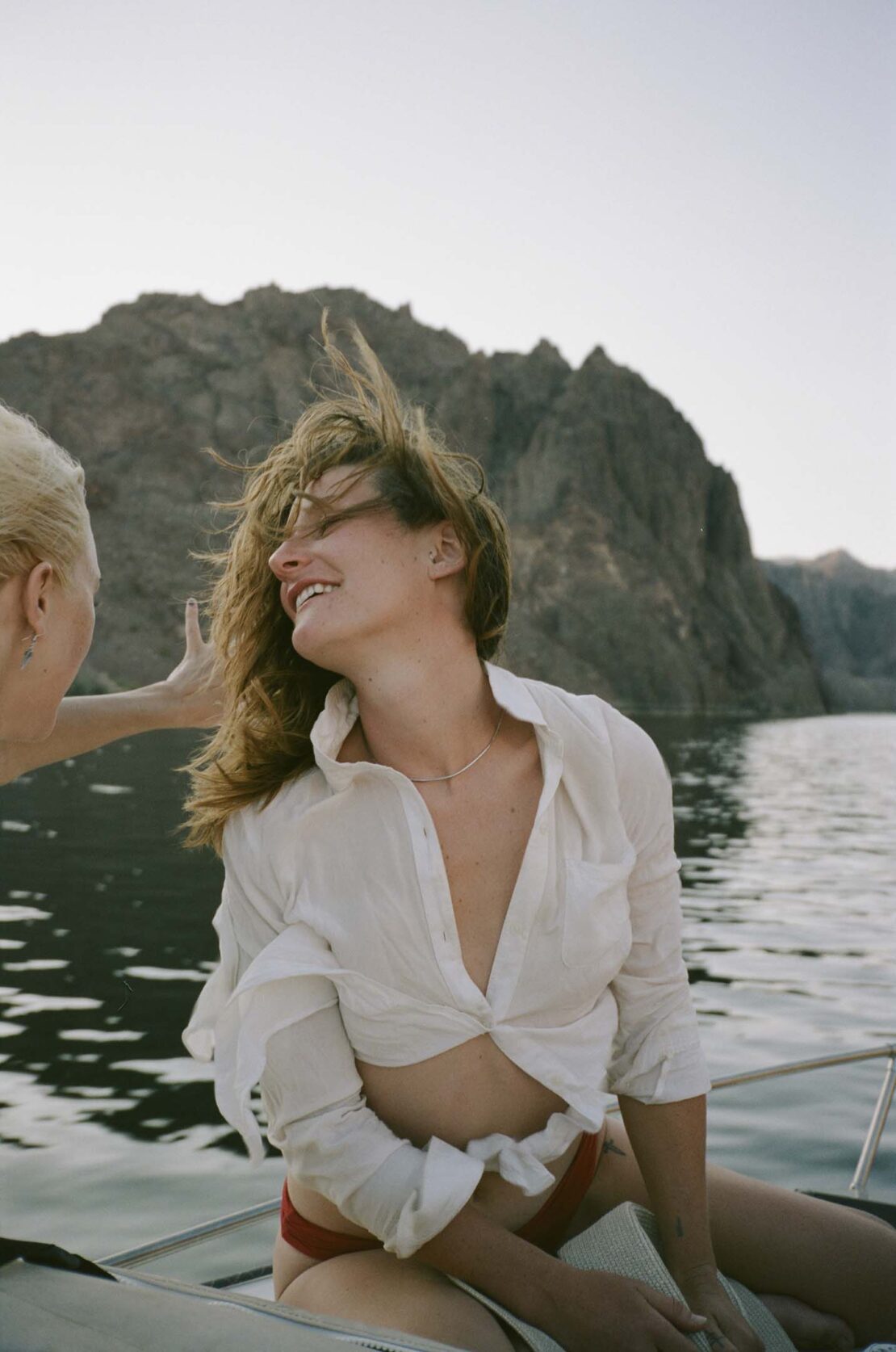 Smiling woman in unbuttoned white shirt sits on boat in water with rocky mountain background; hair blowing in breeze, sunny day.
