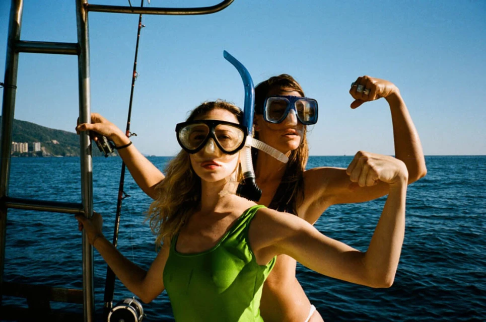 Two women pose on a boat wearing snorkel gear, showcasing swimwear against a backdrop of the ocean, clear blue sky, and coastline.