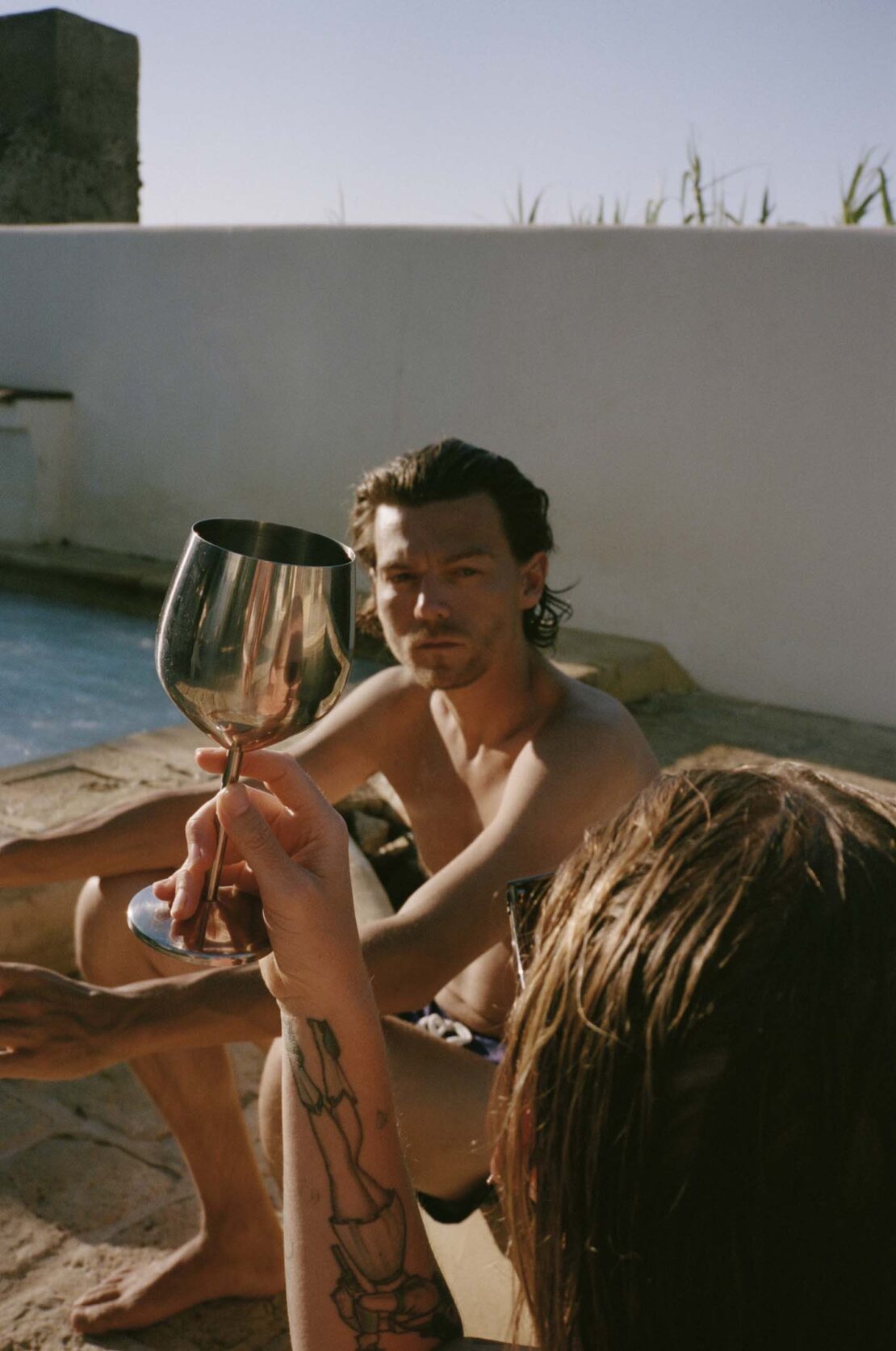 Two people next to a pool with one person raising a wine glass in a sunny outdoor setting, creating a relaxed atmosphere.
