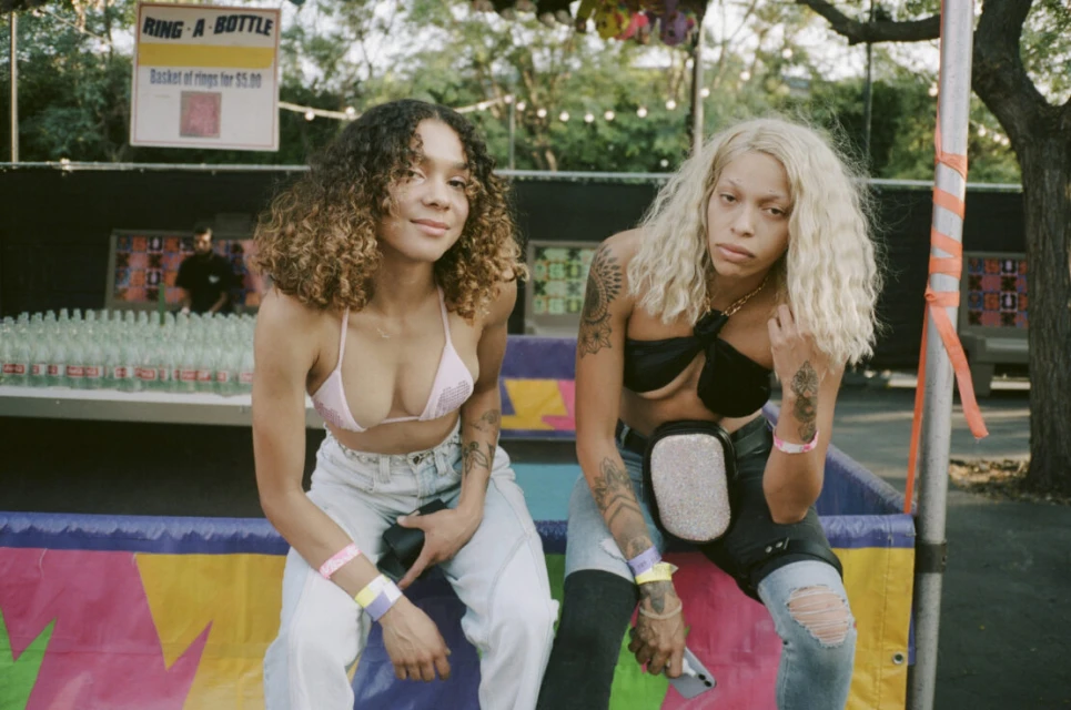Two young women with casual streetwear fashion sit side-by-side at an outdoor event with a booth and string lights in the background.