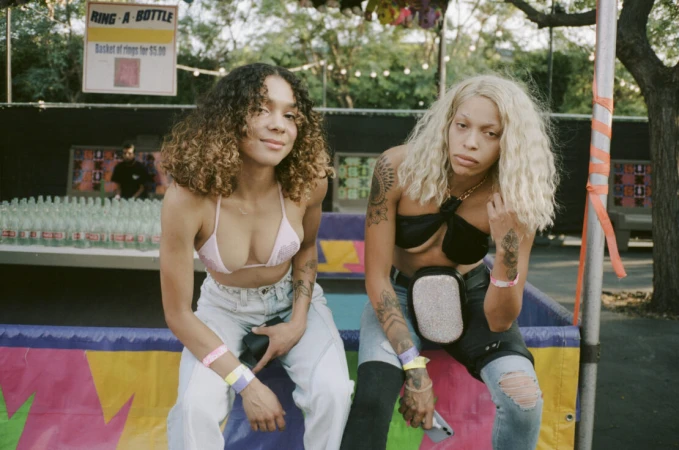Two young women with casual streetwear fashion sit side-by-side at an outdoor event with a booth and string lights in the background.