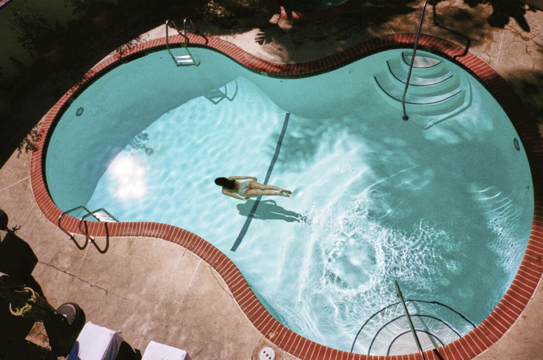 Aerial view of an adult woman floating in a clear blue outdoor swimming pool surrounded by a brick and stone patio and nearby trees.