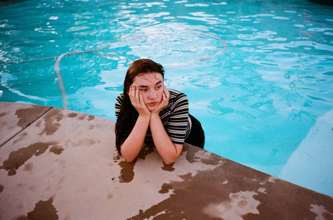 Young woman with wet hair rests chin on her hands while sitting at the edge of a swimming pool on a sunny day.