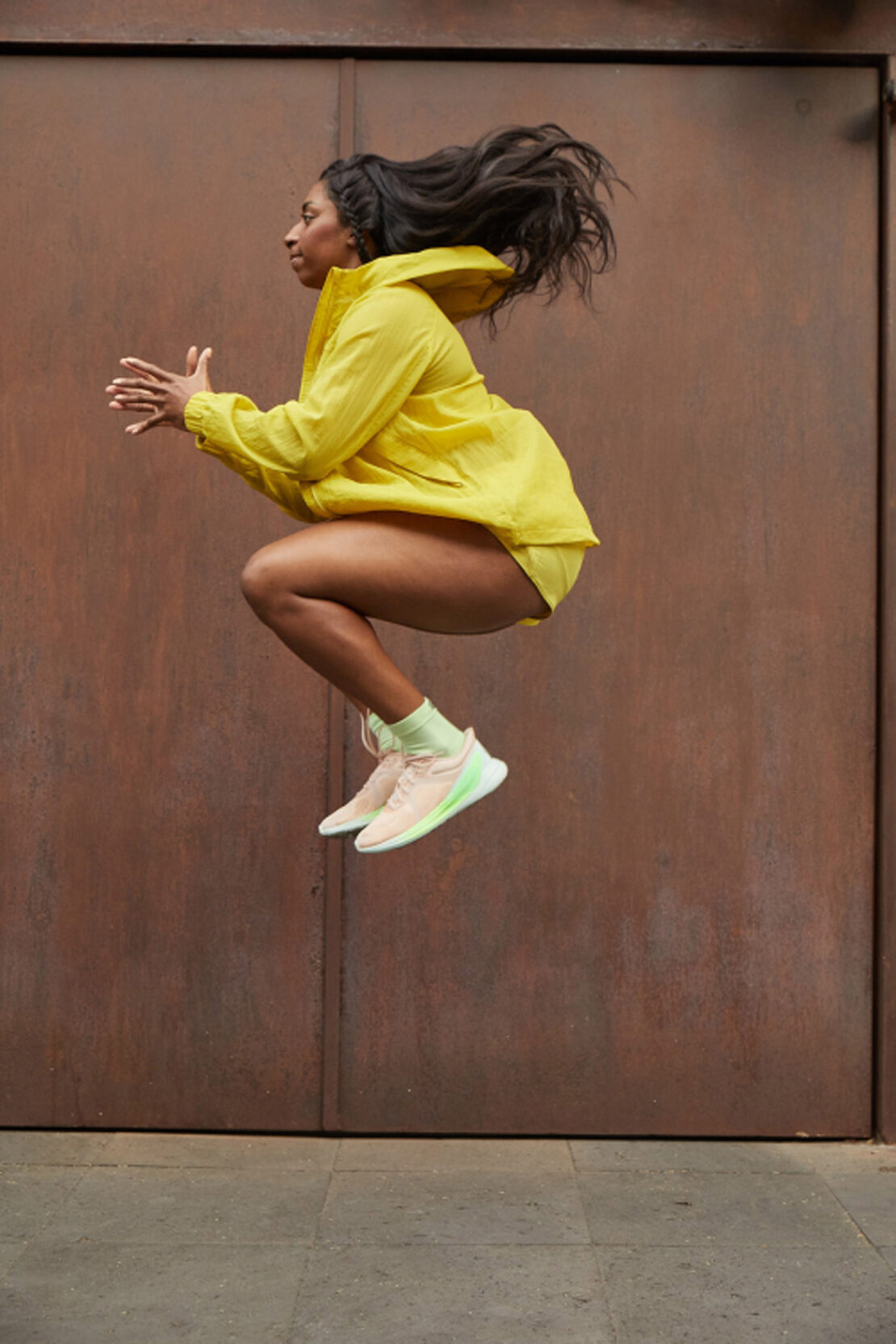 Athletic woman in yellow sportswear dynamically leaps against a textured brown background, showcasing fitness and fashion.