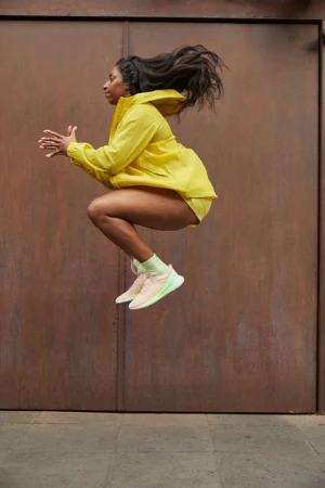 Athletic woman in yellow sportswear dynamically leaps against a textured brown background, showcasing fitness and fashion.